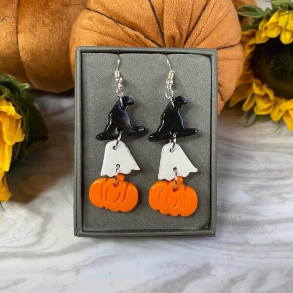 Halloween themed triple dangle earrings with a black hat, white ghost and orange pumpkin handmade polymer clay charms on sterling silver ear wires in a gift box against a velvet pumpkin background from the Janice's Jewels jewellery collection.
